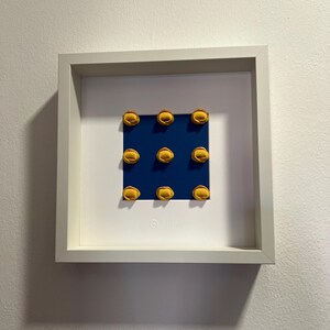 K&ouml;nnte beinhalten: Gerahmtes Kunstwerk mit neun gelben, halbkreisf&ouml;rmigen Objekten, angeordnet in einem 3x3 Raster auf einem dunkelblauen Hintergrund. Das Kunstwerk befindet sich in einem wei&szlig;en quadratischen Rahmen, an einer wei&szlig;en Wand.