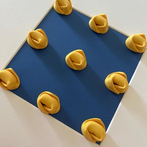 K&ouml;nnte beinhalten: Nahaufnahme von zehn gelben Tortellini auf einem blauen Quadrat, vor wei&szlig;em Hintergrund. Die Pasta hat eine leuchtend gelbe Farbe, und das blaue Quadrat ist von einem wei&szlig;en Rahmen umgeben. Das Bild ist gut beleuchtet und die Pasta ist scharf.