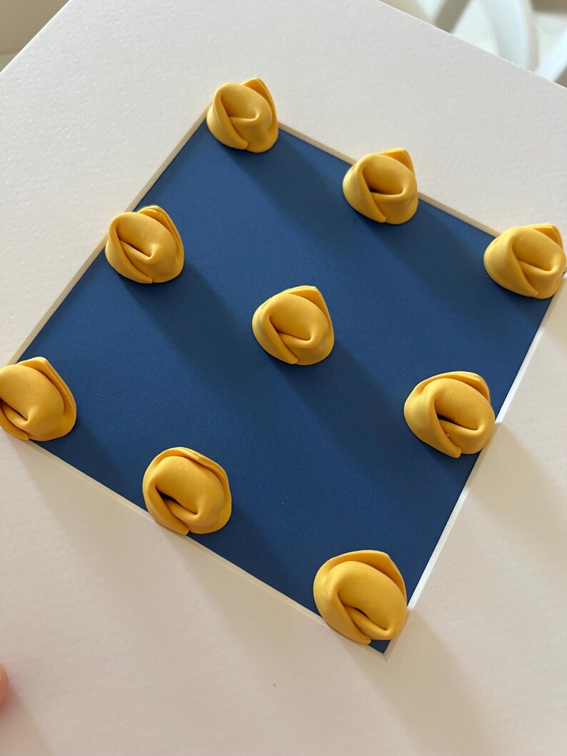 K&ouml;nnte beinhalten: Nahaufnahme von zehn gelben Tortellini auf einem blauen Quadrat, vor wei&szlig;em Hintergrund. Die Pasta hat eine leuchtend gelbe Farbe, und das blaue Quadrat ist von einem wei&szlig;en Rahmen umgeben. Das Bild ist gut beleuchtet und die Pasta ist scharf.