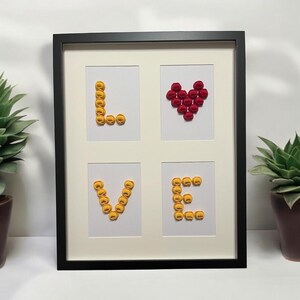 Può includere: Un'opera d'arte incorniciata che presenta la parola "LOVE" scritta con bottoni gialli e un cuore rosso al centro.