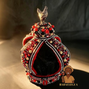 Puede incluir: Una corona con una figura de gallo dorada en la parte superior, sobre una cúpula adornada con cuentas rojas y negras, cristales facetados y detalles dorados. La estructura incluye una base de terciopelo negro con borde rojo y abalorios.