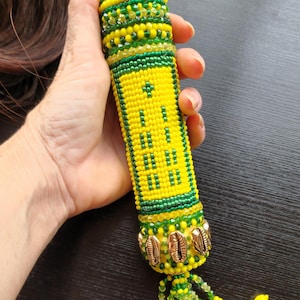 May include: A yellow and green beaded hair wrap with a tassel and cowrie shells. The hair wrap is decorated with a pattern of green and yellow beads.