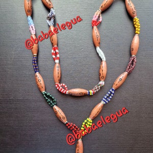 Pode incluir: Um colar longo com contas de madeira castanha alongadas marcadas com símbolos pretos, intercaladas com secções de contas de vidro multicoloridas em amarelo, vermelho, azul, verde e branco. O colar termina numa borla de contas com tons verdes.