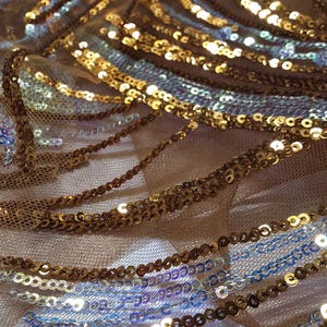 Pode incluir: Close-up de tecido brilhante com lantejoulas douradas, prateadas e azuis. As lantejoulas são costuradas em uma malha bege transparente, criando uma superfície texturizada e elegante. Ideal para roupa de noite ou projetos decorativos.
