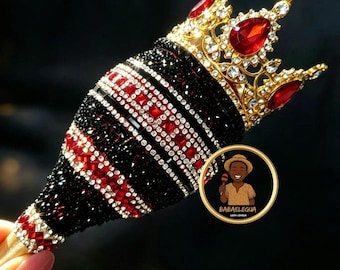 May include: A maraca decorated with black and red rhinestones in a striped pattern, topped with a gold-tone metal crown featuring red teardrop-shaped stones and clear crystals. A circular logo with a cartoon figure and the text BABAELEGUA is visible.