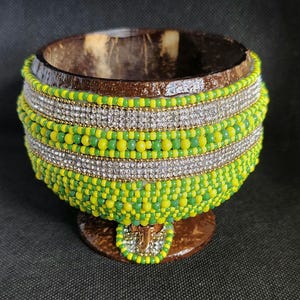 Pode incluir: Uma tigela decorativa feita de casca de coco, adornada com contas verdes e amarelas. Faixas prateadas com detalhes brilhantes circundam a tigela. A tigela repousa sobre uma pequena base redonda.