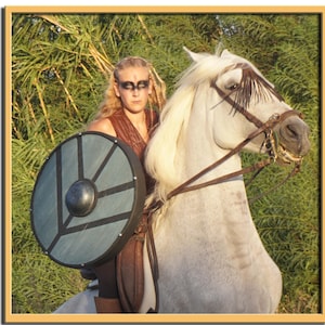 Puede incluir: Una mujer con un chaleco de cuero marrón y un escudo monta un caballo blanco a través de un campo de vegetación verde.