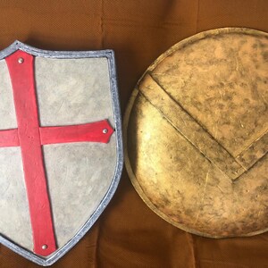 May include: Two shields, one with a red cross on a gray background and the other a round, gold shield with a darker gold design.