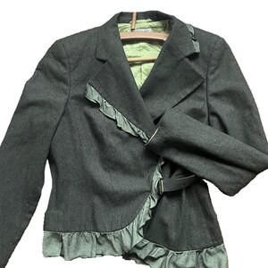 Pode incluir: Um blazer cinza-escuro com forro verde-claro e acabamento em babados verde-claro a condizer ao longo da lapela e da bainha inferior. O casaco inclui um fecho de fivela lateral e mangas compridas, apresentado num cabide de madeira.