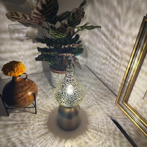 Puede incluir: Una lámpara de mesa dorada con una pantalla estampada, que proyecta intrincados patrones de luz. La lámpara está sobre una mesa junto a una planta en maceta con hojas rayadas y un pequeño jarrón con una flor amarilla.