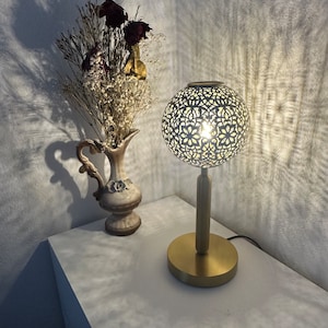 May include: A gold table lamp with a white, intricately patterned lampshade. The lamp is on a white table next to a white ceramic vase with dried flowers.