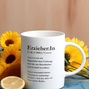 May include: White ceramic mug with a black design that reads "Erzieher:In [e:etsi:ǝbIn] Nomen 1.(n.) ist wie ein Superheld ohne Umhang, der/die mit Bastelkleber und Geduld bewaffnet. 2.(n.) verwandelt kleine Chaoten in zukünftige Genies- und das alles vor dem ersten Kaffee."
