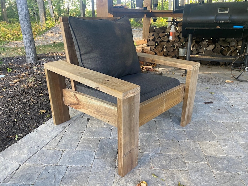 Puede incluir: Una silla de exterior de madera con un coj&iacute;n gris oscuro. La silla tiene un dise&ntilde;o simple y moderno con patas gruesas y robustas.