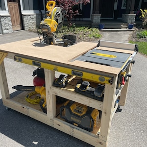May include: A wooden workbench with a yellow and black miter saw, a table saw, and a level. The workbench has two shelves, one with a leaf blower and a red air compressor, and the other with a Dewalt power tool and two batteries.
