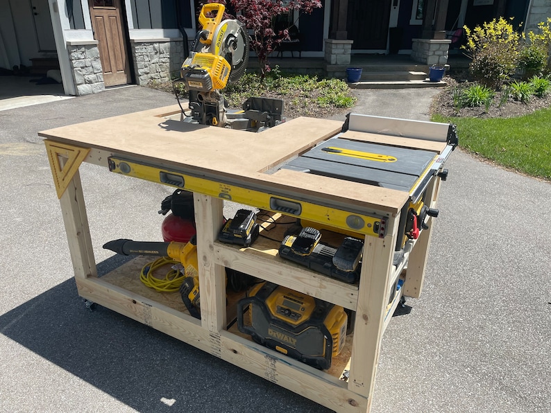 May include: A wooden workbench with a yellow and black miter saw, a table saw, and a level. The workbench has two shelves, one with a leaf blower and a red air compressor, and the other with a Dewalt power tool and two batteries.