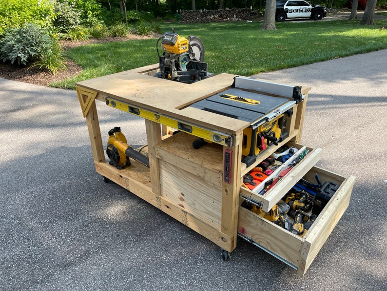 May include: A wooden workbench with a yellow and black table saw, a yellow and black miter saw, and a yellow leaf blower. The workbench has two drawers that are open and filled with tools. The workbench is on wheels and is in a driveway.