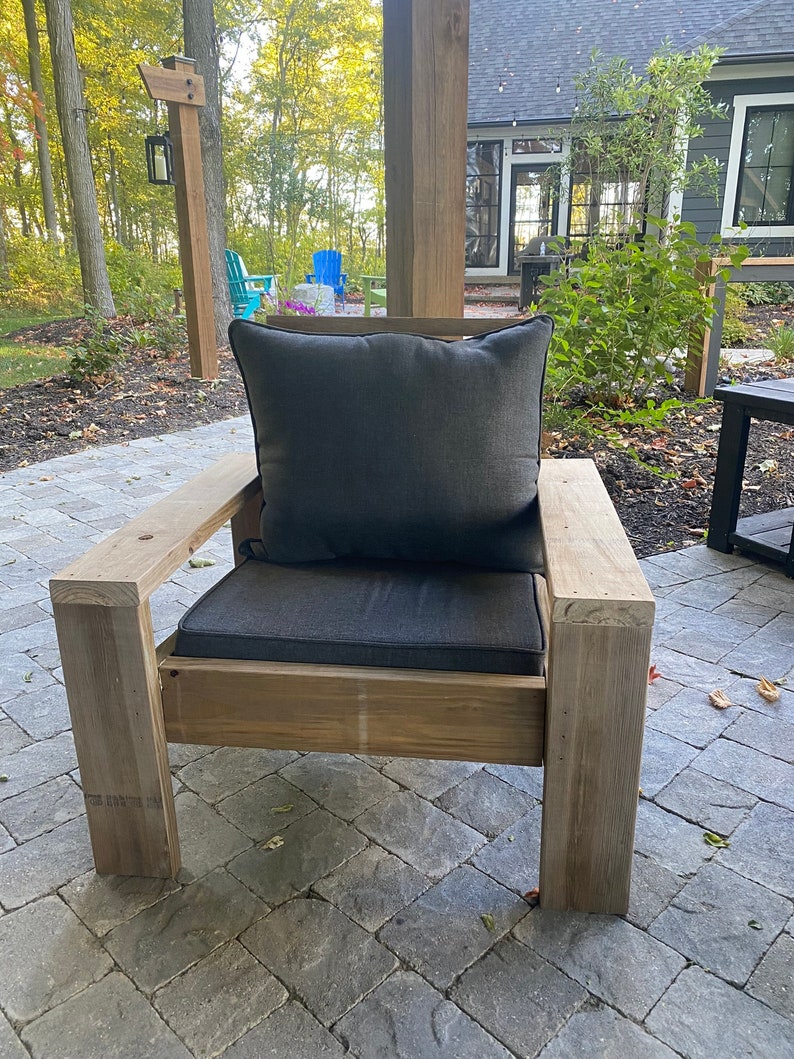 Puede incluir: Una silla de madera con un coj&iacute;n gris y una almohada gris. La silla est&aacute; en un patio con suelo de ladrillo.