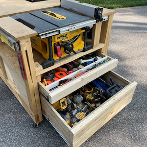 May include: A wooden workbench with a yellow and black table saw mounted on top. The workbench has two drawers, one open and one closed, filled with tools. The workbench is on wheels and is sitting on a paved driveway.