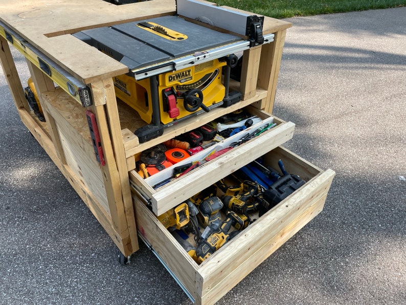 May include: A wooden workbench with a yellow and black table saw mounted on top. The workbench has two drawers, one open and one closed, filled with tools. The workbench is on wheels and is sitting on a paved driveway.