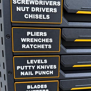 May include: A dark grey tool chest drawer front featuring rectangular labels with orange borders and white text. The labels are organised by category: SCREWDRIVERS NUT DRIVERS CHISELS, PLIERS WRENCHES RATCHETS, LEVELS PUTTY KNIVES NAIL PUNCH, and BLADES CUTTERS PENCILS.