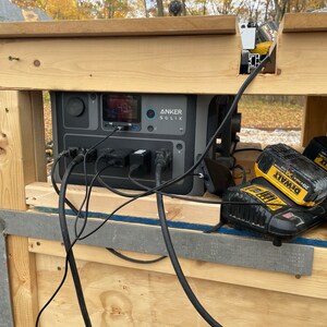 May include: A black portable power station with the text "ANKER SOLIX" on the front. The power station is plugged into a wooden table with multiple cords and two yellow and black DeWalt batteries.