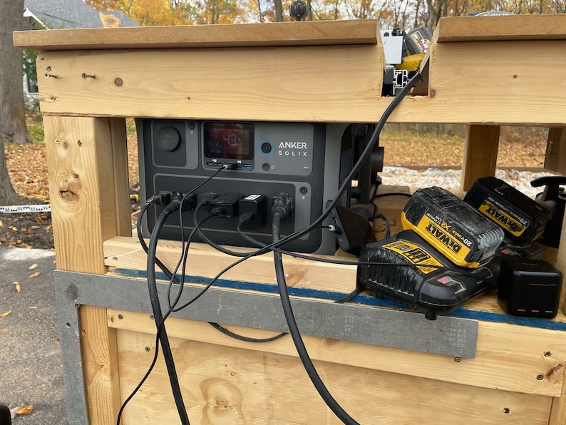 May include: A black portable power station with the text "ANKER SOLIX" on the front. The power station is plugged into a wooden table with multiple cords and two yellow and black DeWalt batteries.