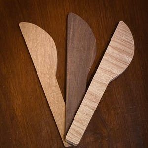 May include: Three wooden butter knives in varying shades of brown. The knives are arranged in a fan shape, with one in a dark brown, one in a medium brown, and one in a light brown. The knives have a curved blade and a long handle.