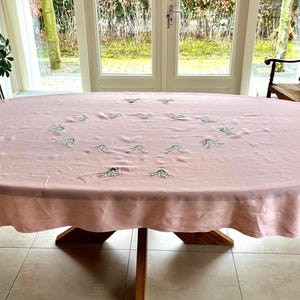 Puede incluir: Un mantel grande, ovalado, de color rosa claro con diseños florales bordados. El mantel cubre una mesa de madera con una base central. La habitación tiene grandes ventanas y una silla de madera.