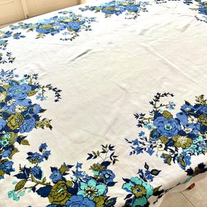 Può includere: Tovaglia bianca con motivo floreale. Il motivo presenta fiori blu, turchese e verde oliva disposti a grappoli sui bordi. Il tessuto sembra morbido, forse lino. Adatta per un tavolo rotondo o ovale.