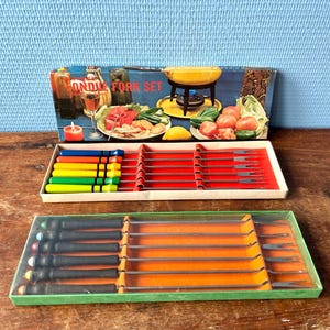 Op de afbeelding: Vintage fonduevorkenset met kleurrijke handvatten en een doos met een illustratie van een fonduepan. De set bevat meerdere vorken in een rode en oranje bak, perfect voor een retro diner. Op de doos staat "Fondue Fork Set".