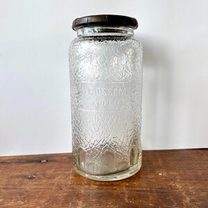 Op de afbeelding: Een heldere glazen pot met een getextureerd oppervlak en een donkerbruin deksel. De pot heeft de tekst "V. ROSSEM. KOFFIE." De pot staat op een houten oppervlak.