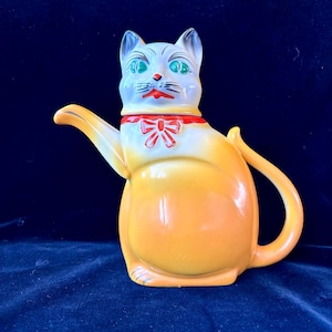 Könnte beinhalten: Eine Keramik-Teekanne in Form einer Katze. Die Katze hat einen weißen Kopf mit blauen Ohren, grünen Augen und einer roten Fliege. Der Körper ist gelb mit einem gebogenen Griff und Ausguss.