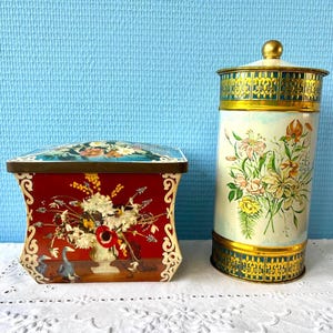 Può includere: Due contenitori in metallo vintage. Uno è una scatola rettangolare con sfondo rosso e design floreale. L'altro è una latta cilindrica alta con design floreale su sfondo crema. Entrambi hanno dettagli dorati.