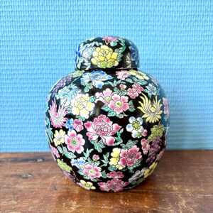 May include: A black ceramic jar with a lid, adorned with a vibrant floral pattern in pink, yellow, blue, and white. The jar sits on a wooden surface against a light blue backdrop. The floral design covers the entire jar and lid.