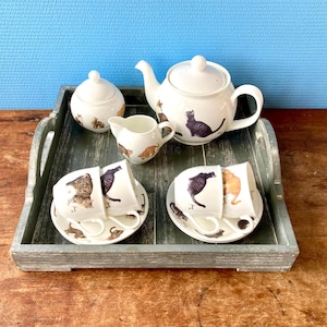 May include: A grey wooden tray with a white teapot, sugar bowl, creamer, and four teacups with cat illustrations. The teacups are arranged in two pairs on the tray.