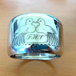 Puede incluir: Un servilletero plateado con un diseño grabado de dos palomas enfrentadas, formando un corazón. Las iniciales "F.H.V." están grabadas debajo. El anillo tiene una superficie pulida y reflectante.