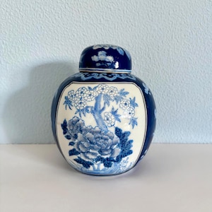 May include: A blue and white ceramic ginger jar with a lid. The jar features a floral design with a large peony and tree branches. The lid has a matching floral pattern. The jar is round with a glossy finish.