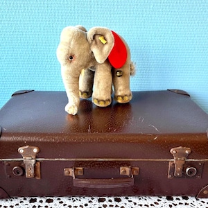 Könnte beinhalten: Ein brauner Vintage-Koffer mit Messingbeschlägen und einem Stoffelefanten mit rotem Ohranhänger. Der Elefant ist hellgrau mit gelben Akzenten und steht auf dem Koffer vor blauem Hintergrund.