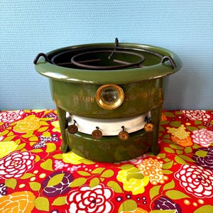 Könnte beinhalten: Ein vintage olivgrüner emaillierter Metallkocher mit einem kreisförmigen oberen Rost. Die Vorderseite verfügt über ein kleines Sichtfenster aus Glas, einen weißen Brennerbereich und vier kleine Metallknöpfe. Der Text BECCON DOETINCHEM ist aufgedruckt.