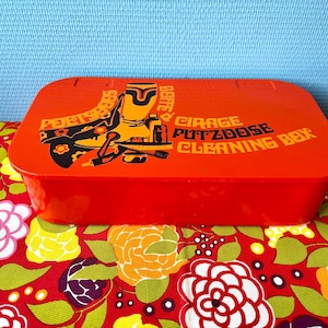 May include: A rectangular, bright orange box with a lid featuring the words "CIRAGE PUTZDOSE CLEANING BOX" in yellow and black text. The box has a graphic of a shoe and cleaning tools. The box sits on a floral patterned fabric.