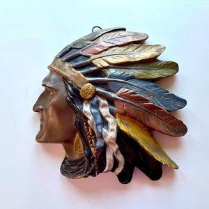 Pode incluir: Uma decoração de parede em tons de bronze que retrata um chefe nativo americano em perfil. A peça apresenta um cocar detalhado com penas em tons de ouro, azul, castanho e verde. A escultura tem uma argola para pendurar.