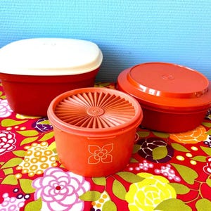 Op de afbeelding: Drie vintage plastic voedselopslagcontainers. De containers zijn rood en oranje met witte en oranje deksels. De containers staan op een tafelkleed met bloemenpatroon met een blauwe achtergrond.