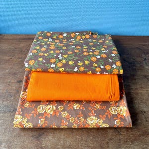 Peut inclure: Trois pièces de tissu pliées. Deux sont marron avec des motifs floraux orange, jaune et blanc. La pièce du milieu est orange uni. Les tissus sont empilés sur une surface en bois avec un fond bleu.