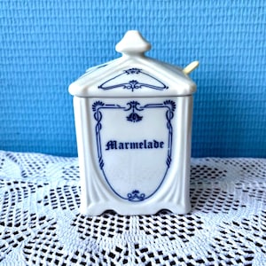 Peut inclure: Un pot à marmelade en céramique blanche avec une base carrée et un couvercle assorti. Le pot présente des motifs décoratifs bleus en volutes et le mot "Marmelade" imprimé en bleu foncé dans une police gothique sur le devant. Une petite cuillère jaune est visible.