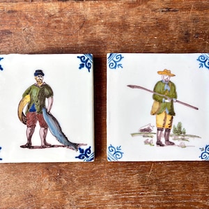 Può includere: Due piastrelle quadrate in ceramica con illustrazioni dipinte a mano. Una raffigura un uomo che tiene un pesce, l'altra un uomo con un bastone. Entrambe hanno disegni blu agli angoli e sono ambientate su uno sfondo di legno.