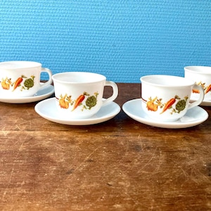 Op de afbeelding: Vier witte theekopjes met een bloemenpatroon van wortels, bladeren en bloemen op een houten oppervlak. Elk theekopje heeft een bijpassend schoteltje.