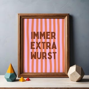 Può includere: Stampa con cornice in legno con le parole "IMMER EXTRA WURST" in marrone su sfondo a righe rosa e arancioni. Forme geometriche decorative gialle, verde acqua e rosse sul tavolo.