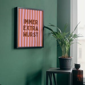 May include: A framed art print with a black border. The print features vertical stripes in shades of pink and orange, with the words "IMMER EXTRA WURST" in brown lettering. The artwork is displayed on a green wall.