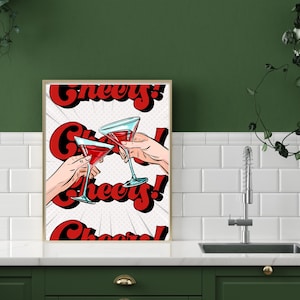 May include: A framed pop art print featuring two hands toasting with red cocktails in martini glasses. The word "Cheers!" is written in red, stylized text. The background is a white and gray dotted pattern.
