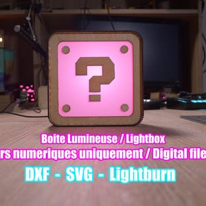 Peut inclure: Une boîte lumineuse carrée avec un cadre en bois et un centre rose éclairé avec un point d'interrogation marron. Le texte "Boite Lumineuse / Lightbox" et "DXF - SVG - Lightburn" sont visibles. L'objet est sur une surface en bois.